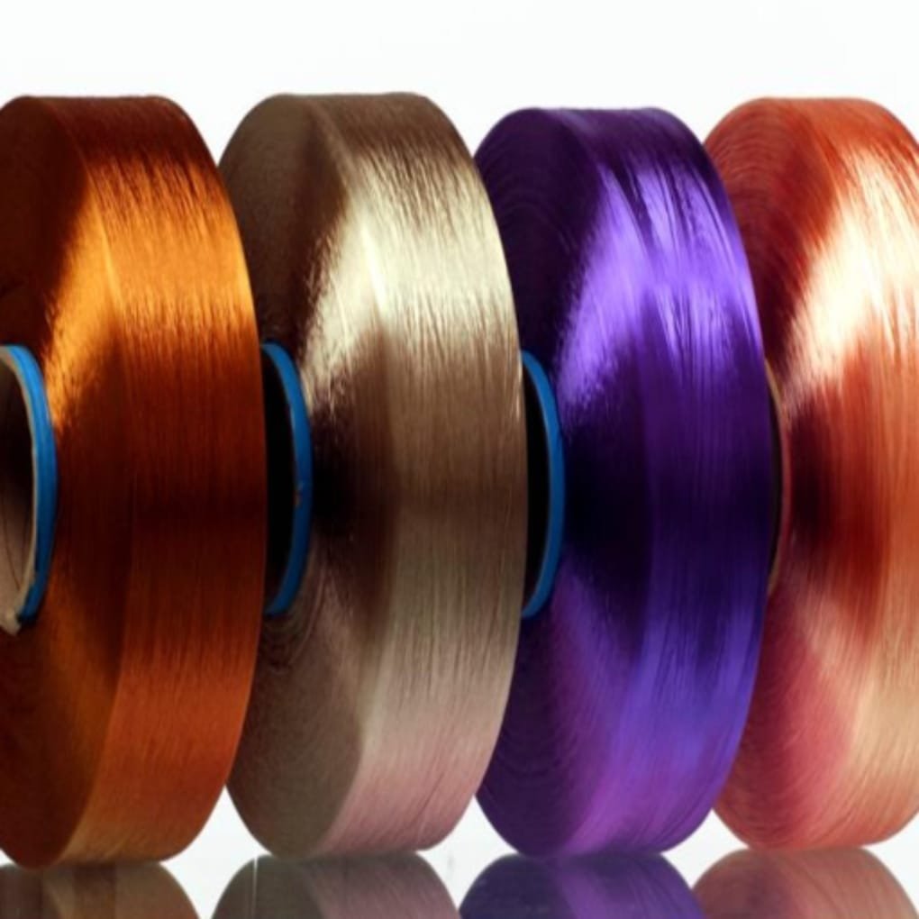 A row of colorful spools of shiny filament or ribbon in orange, brown, gold, purple, and red, lined up in a straight row with reflections below.