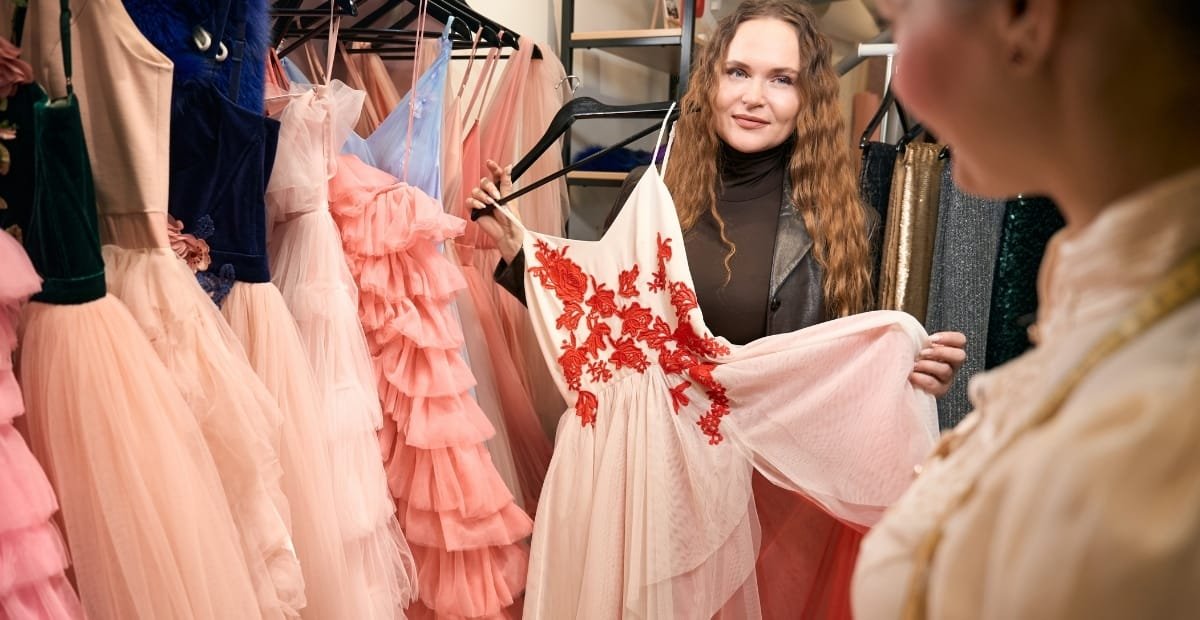 Haute Couture Fashion: Where Artistry Meets Luxury Design A smiling designer in a boutique holds up a pale pink gown with bold red floral embroidery, surrounded by racks of pastel-hued, ruffled dresses.