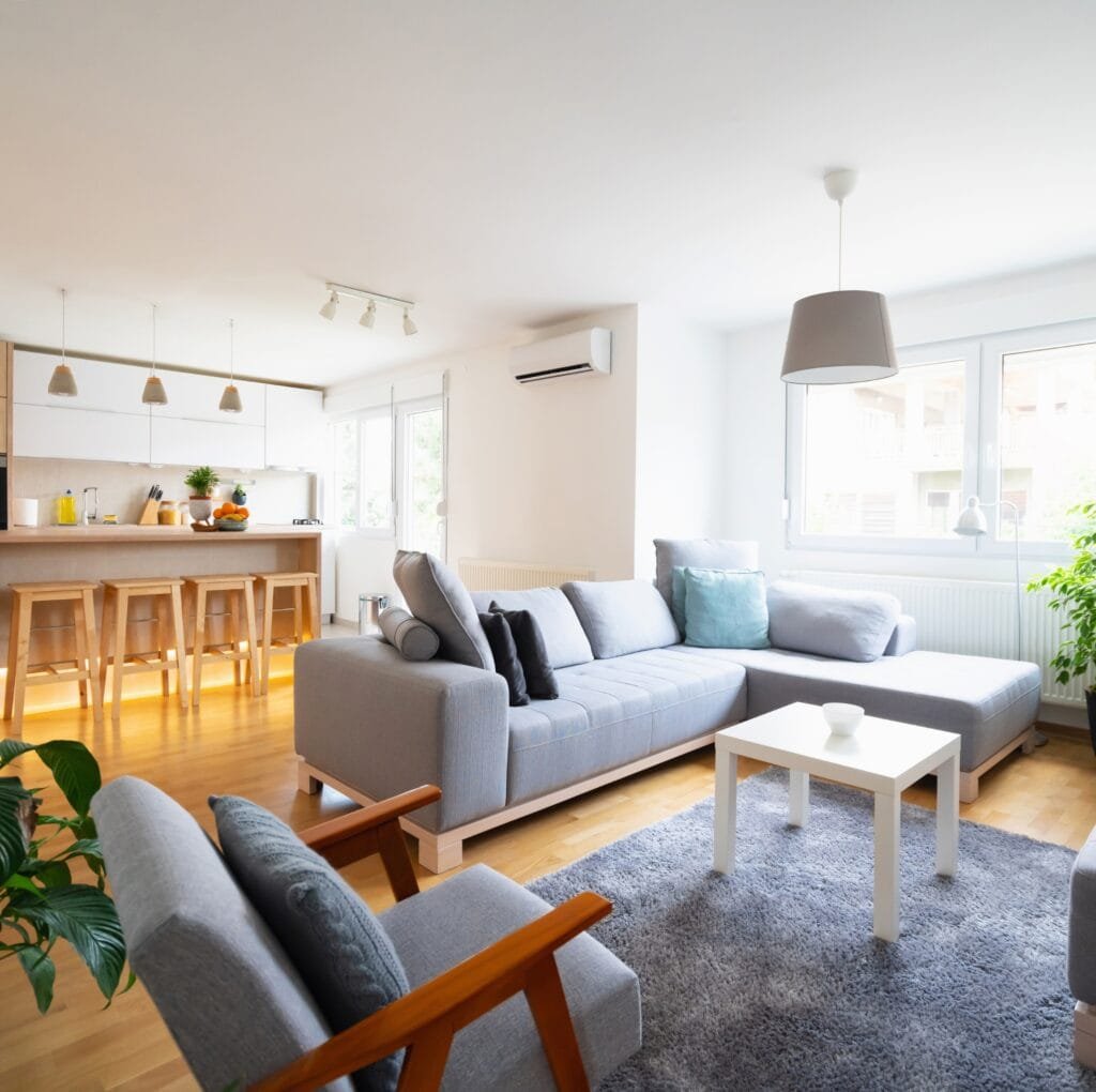 Modern open-plan living room with a light gray sectional sofa, matching lounge chairs, a white coffee table, and a soft blue-gray area rug. The kitchen island with wooden stools is in the background. Large windows bring in natural light, and greenery sits by the window. Neutral tones and clean lines create a calm, contemporary aesthetic.
