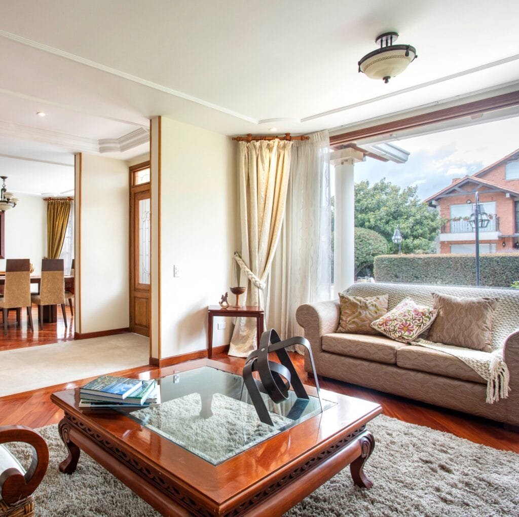A bright, elegant living room featuring a cream-colored sofa with patterned throw pillows, a glass-top wooden coffee table, and a shaggy area rug. Large windows with sheer linen curtains overlook a suburban street. A dining area with a wooden table and upholstered chairs is visible in the background. The room has warm wood floors, decorative molding, and soft natural light.