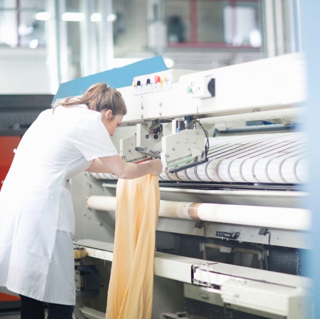 An industrious worker in a white lab coat operates a modern textile loom, guiding a soft, pale-yellow linen fabric as it runs through the machine in a bright, clean factory setting.