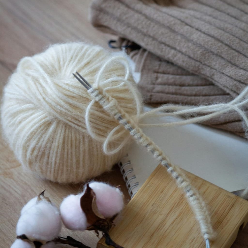 A cozy knitting setup: a ball of cream-colored yarn with a knitted swatch in progress on double-pointed needles, plus a cotton plant branch with brown pods and pink-tinted cotton bolls in the foreground. A striped beige textile and a small notebook sit in the background.

