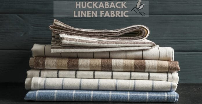 A neatly stacked pile of folded linen fabrics in earthy, natural tones (beige, tan, cream, and light blue) with subtle stripe patterns. The top of the image shows a header reading “Huckaback Linen Fabric” with a small scissors illustration.