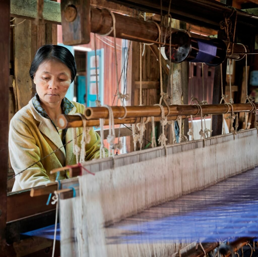 A woman weaving on a traditional loom with wooden beams and hanging cords in a rustic workshop.