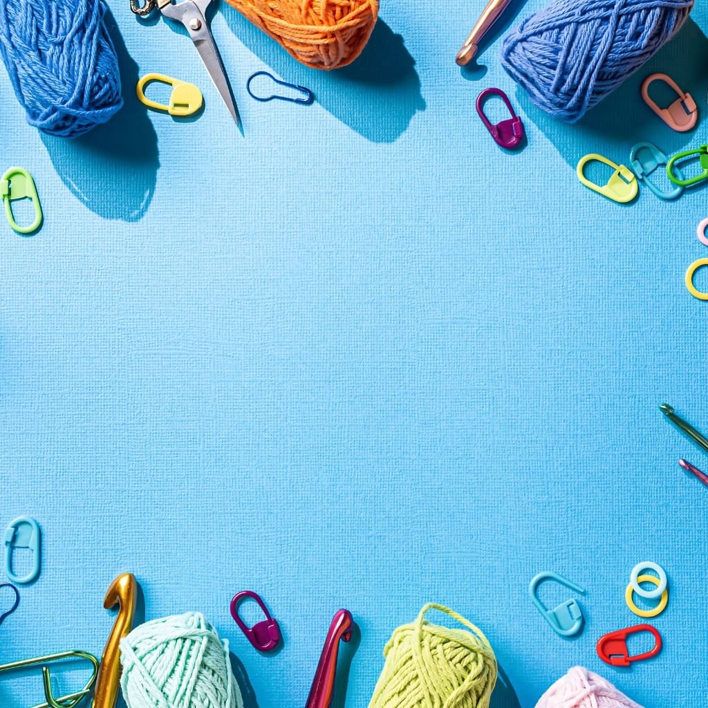 A bright blue knitting workspace bordered with colorful yarn balls, crochet scissors, and assorted plastic stitch markers and knitting tools. The center is open for focus on a knitting project.