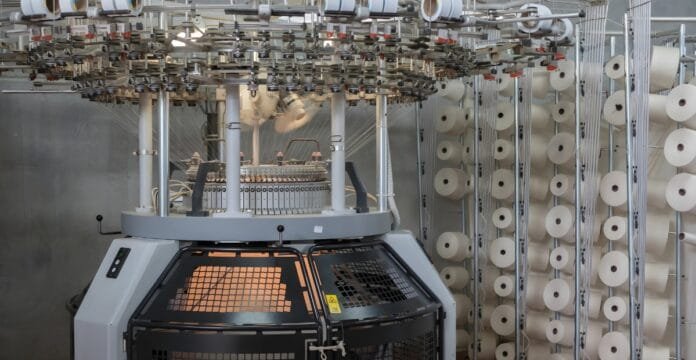 A high-tech circular knitting machine in a textile factory, surrounded by numerous white spools of thread and threading components. The machine has a protective mesh door and glowing orange interior, with many needles and yarn guides overhead.