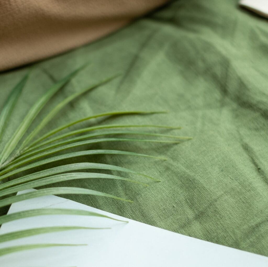 Close-up of a green linen fabric with a palm leaf placed on top, highlighting natural textures and a soft, earthy color palette.