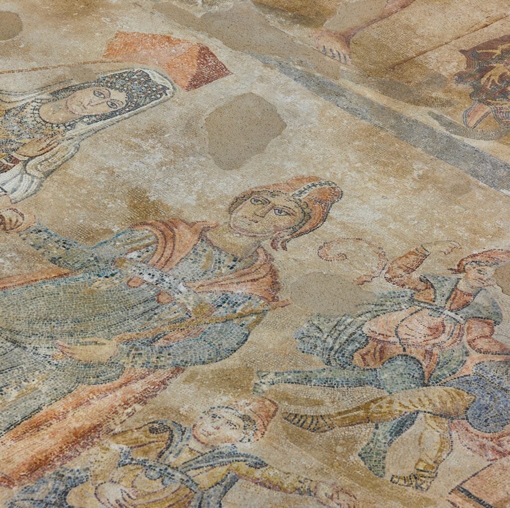 A medieval linen fabric motif shows a mosaic-like scene with stylized human figures in armor and flowing garments, rendered in muted earth tones. The image resembles a fragment of a wall or floor mosaic from the Middle Ages, with worn details and pale colors.
