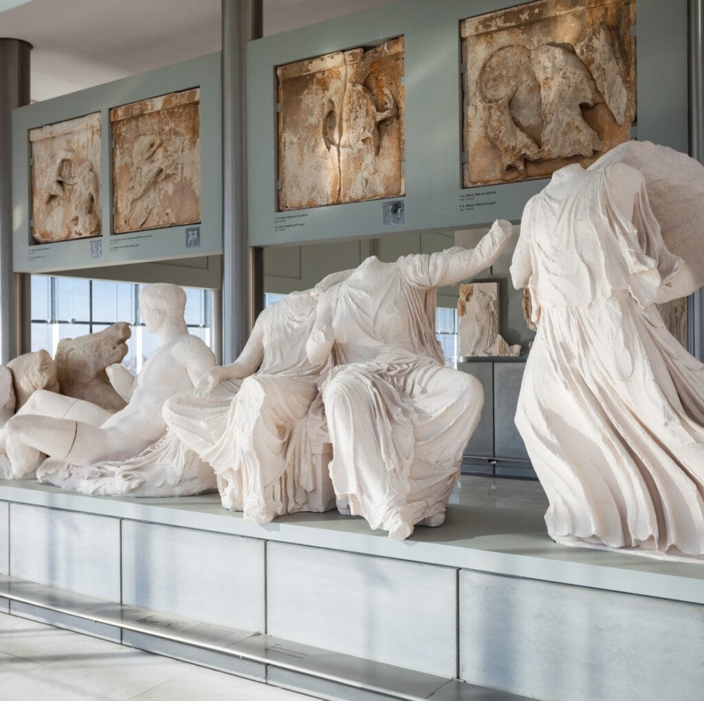 A row of white plaster statues draped in flowing garments sits on a display platform in a museum, with weathered stone reliefs mounted in display cases overhead.
