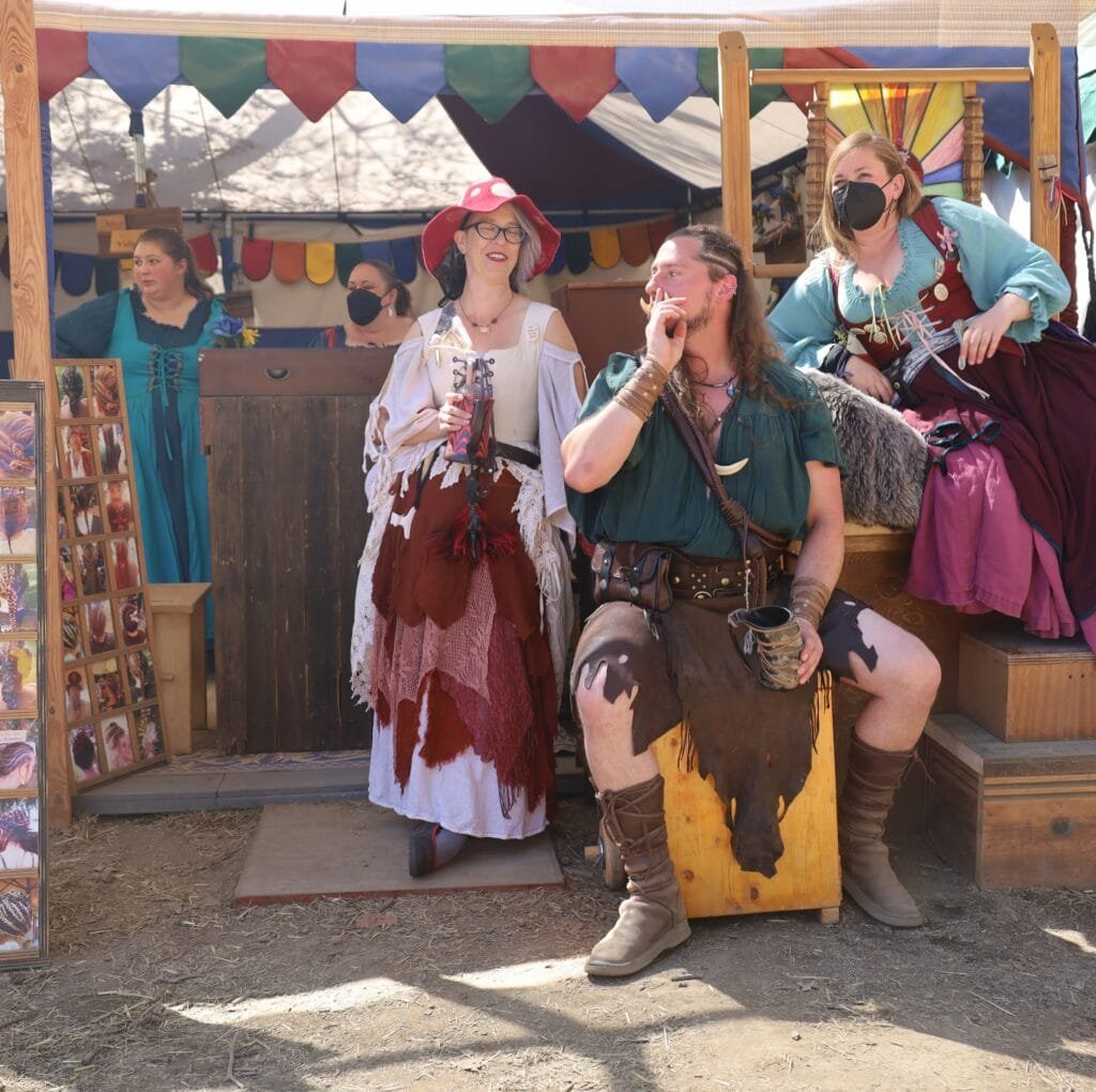 A group of people in Renaissance-style costumes at a fair: a man in a green tunic and brown leather gear sits on a wooden block, smoking and holding a mug; a woman in a red-and-white layered dress with a white shawl and red hat stands beside him; two other costumed women, one in blue-green gown and the other in a teal-gray gown with a mask, stand behind a wooden booth decorated with colorful pennant banners. The ground is dirt, and there are display boards and photos around the booth.