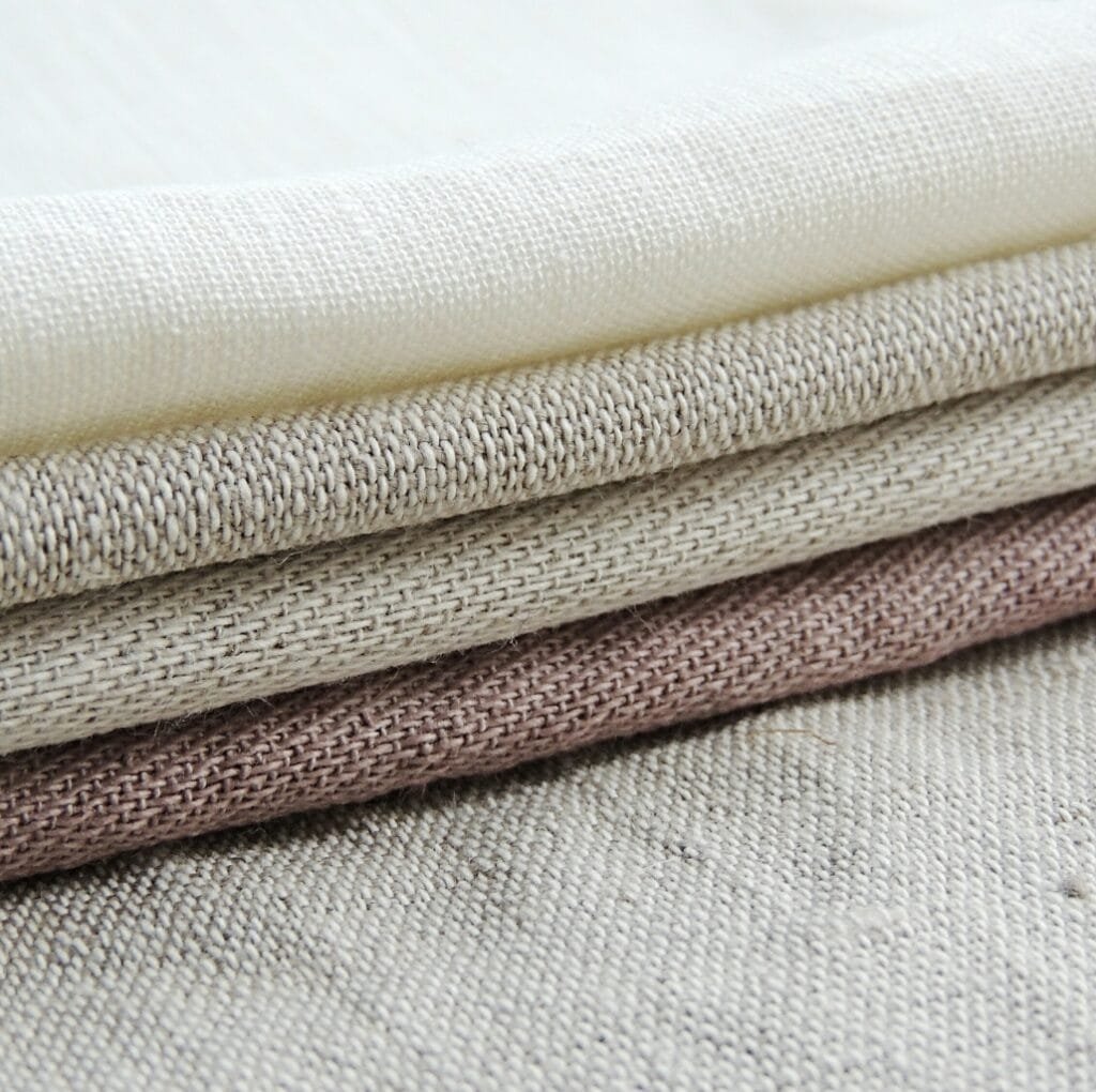 A stack of folded linen fabric with loose, open weaves in neutral tones (cream, beige, and light brown).