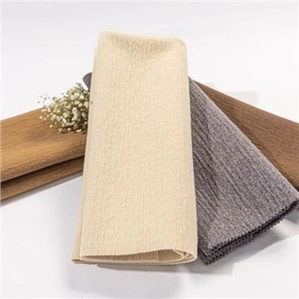 A trio of folded fabric samples: a light beige textured fabric in the foreground, a gray textured fabric behind it, and a wooden plank with small white flowers in the background.