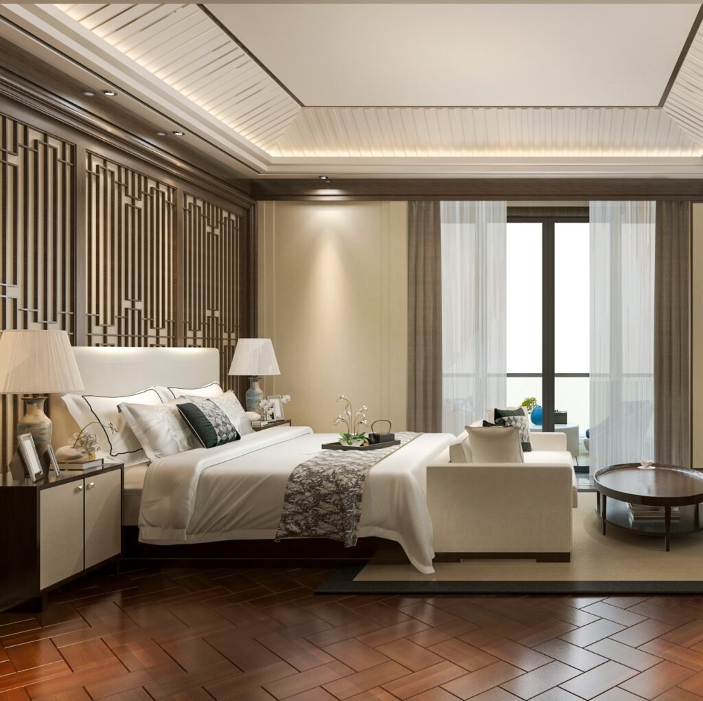 A luxurious hotel-style bedroom featuring a large bed with white linens and decorative damask-patterned throw, dark wood nightstands, elegant table lamps, and a seating area near floor-to-ceiling windows with sheer curtains; the rich herringbone wooden floor adds warmth to the modern decor.