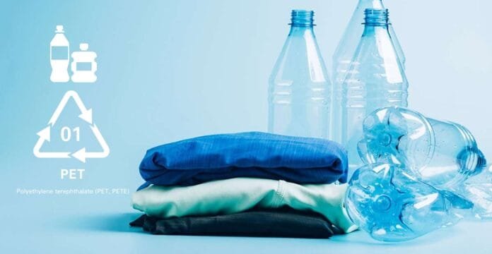 A light blue background with folded clothes in navy, teal, and black stacked in the foreground. To the left, white recycling symbols and plastics icons appear. In the background, several clear plastic bottles and a crushed bottle lay on the right side.
