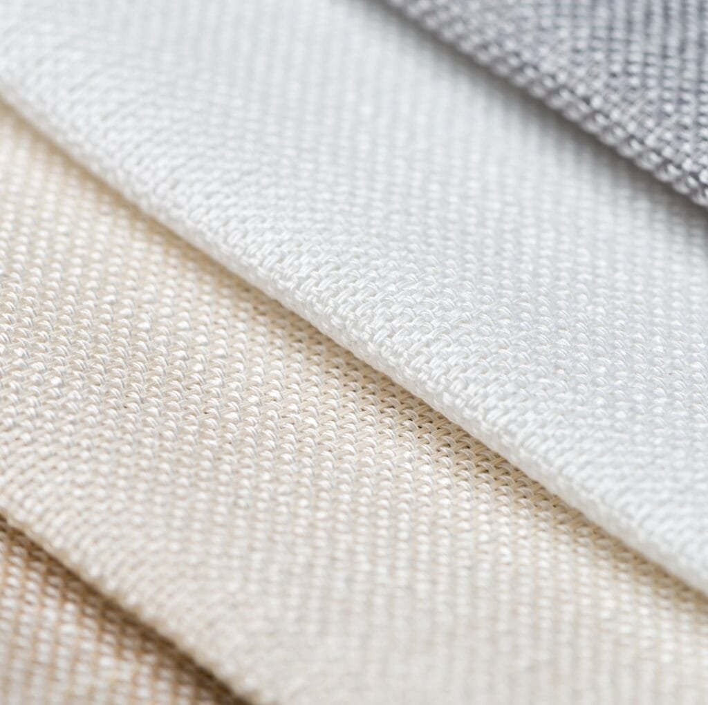 Close-up image of three layers of plain weave linen fabric in shades of beige, white, and gray, showcasing the woven texture and detailed interlacing of the threads.