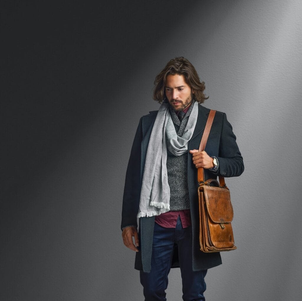 A stylish man stands against a neutral gray backdrop wearing a formal coat and scarf, carrying a brown leather satchel over his shoulder.