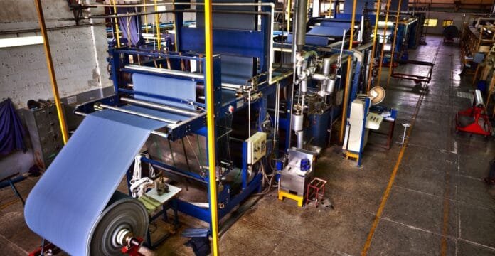 An industrial textile production floor with large blue fabric sheets on a conveyor system, surrounded by heavy machinery, rollers, and metal frames.