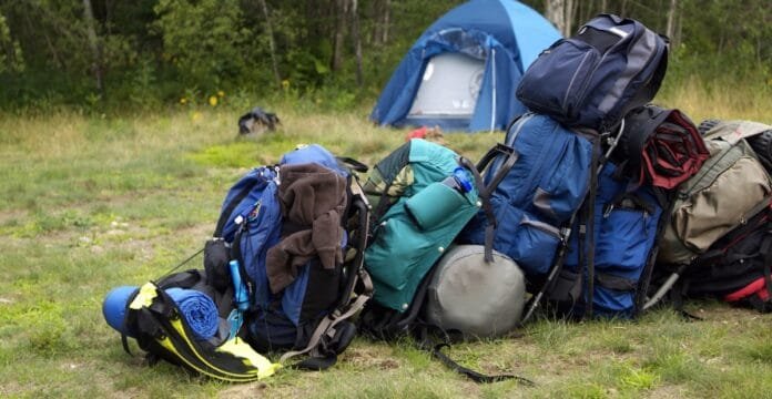 A pile of colorful outdoor backpacks and gear stacked on a grassy campsite, with a blue tent in the background.