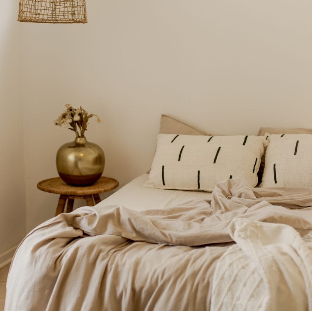 A neatly made bed with beige sheeting linen fabric, featuring wrinkled and slightly disheveled sheets and pillows. A small wooden nightstand with a gold vase and dried flowers sits beside the bed, and a woven lampshade hangs overhead.