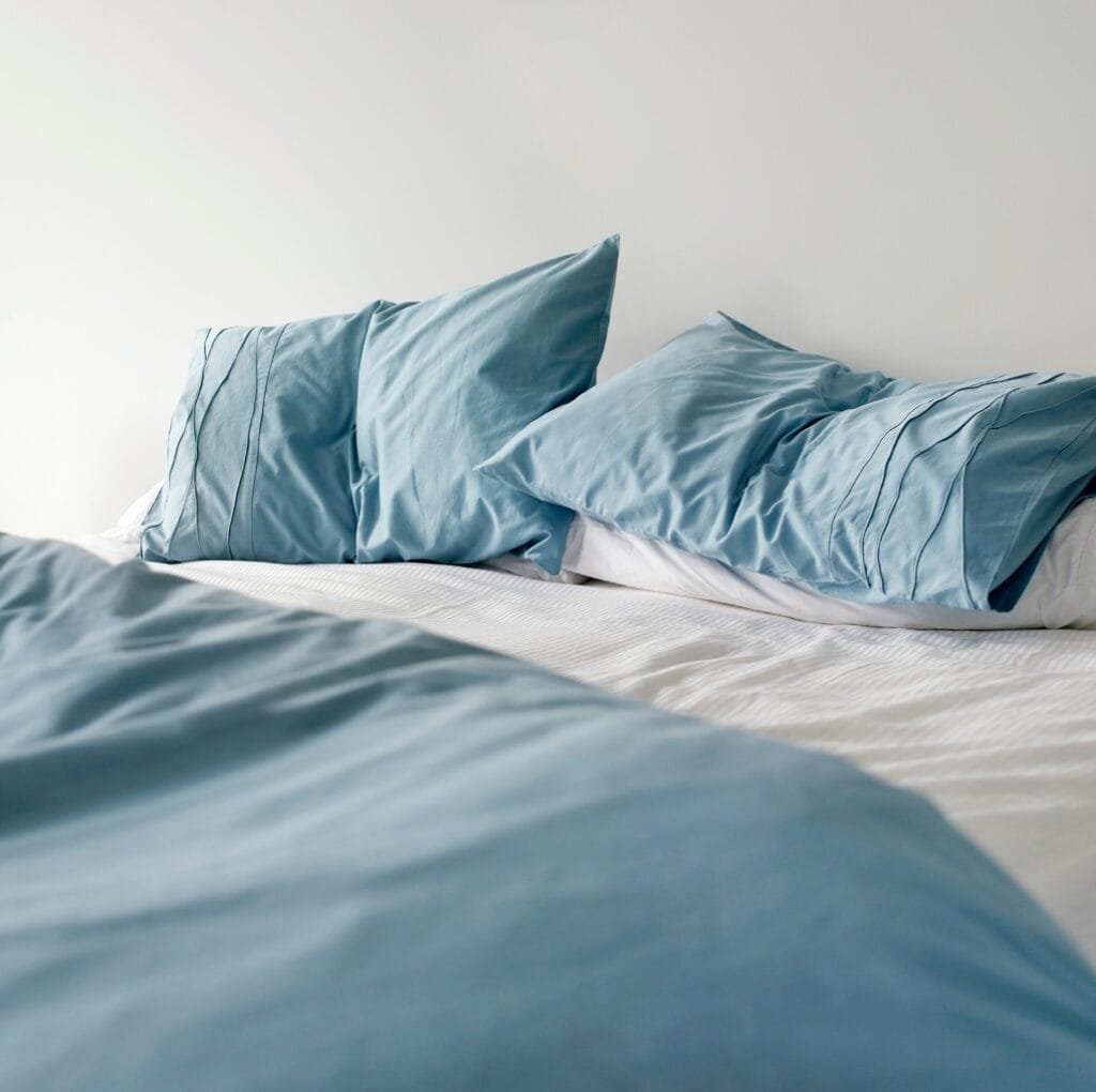 A neatly made bed with soft blue linen sheets and matching blue pillowcases. The sheets have a light, airy texture and gentle folds, set against a plain white wall.