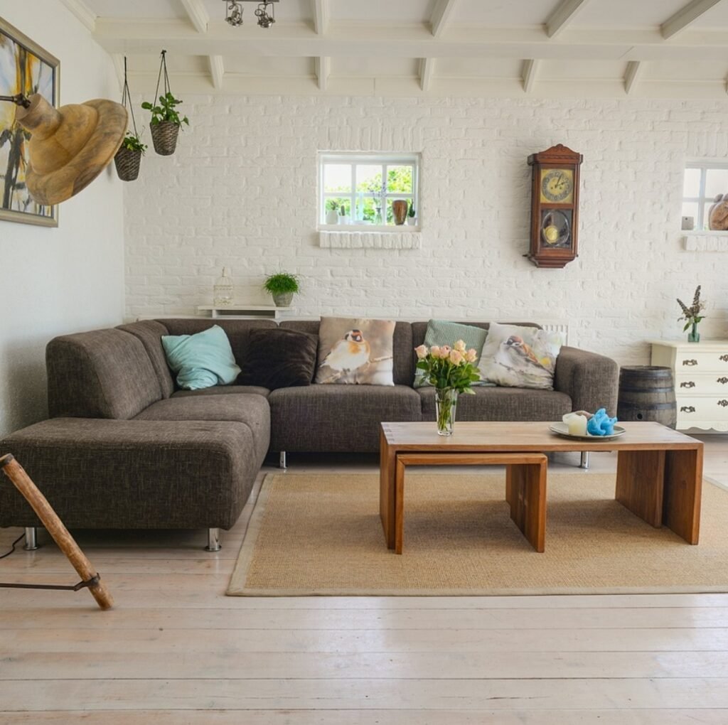 A bright, cozy living room with a large dark brown sectional sofa, a light wooden coffee table, and a natural fiber rug. White brick walls, small windows, and a vintage wall clock add charm. Green plants and soft pastel pillows complete the décor.