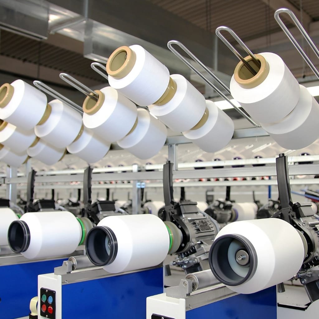 Close-up view of spinning machinery in a cotton mill, with spinning rollers and fibers feeding into the spool, highlighting the mechanical process of turning cotton fibers into yarn.