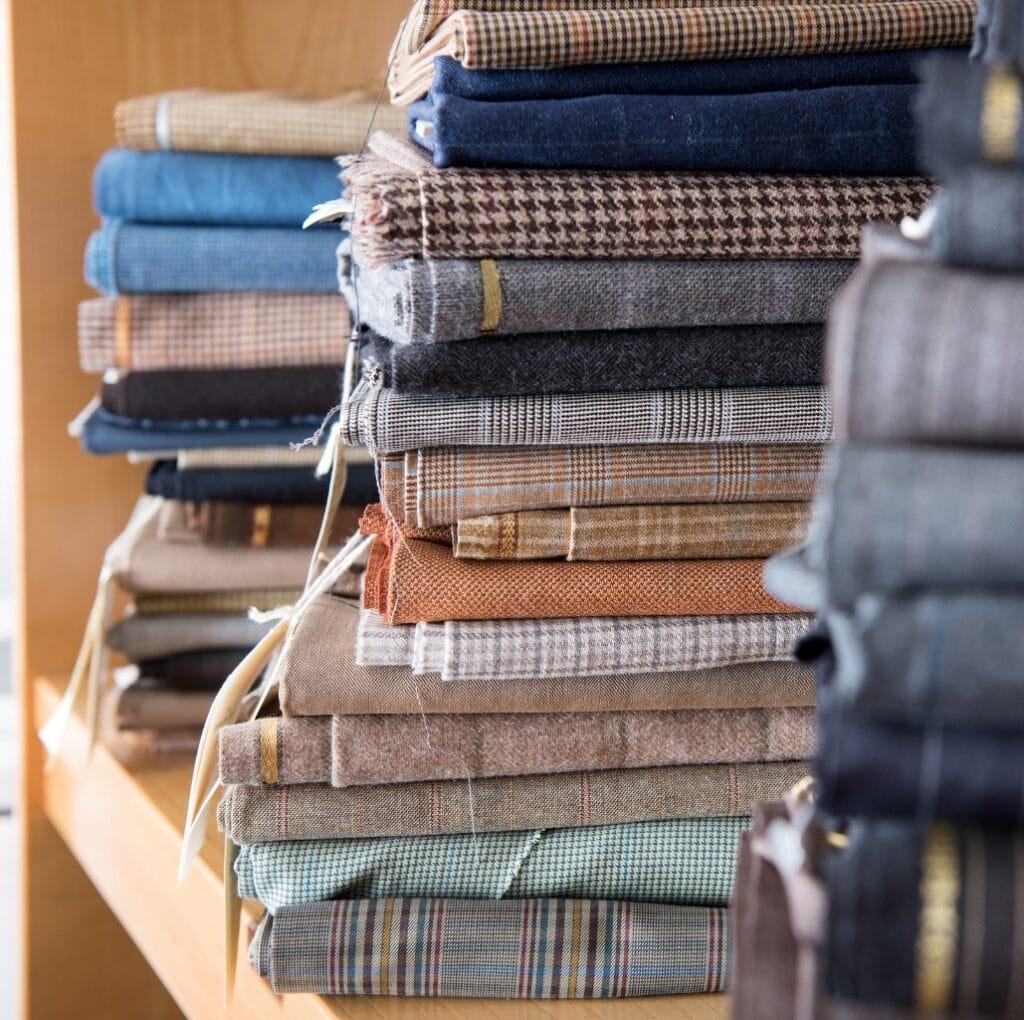 A tall stack of folded suiting fabrics in a fabric shop. The rolls feature a variety of textures and patterns, including houndstooth, plaids, tweeds, and solid weaves in earthy browns, blues, and grays. The fabrics are neatly piled on wooden shelves with some light fringing and small tags visible.