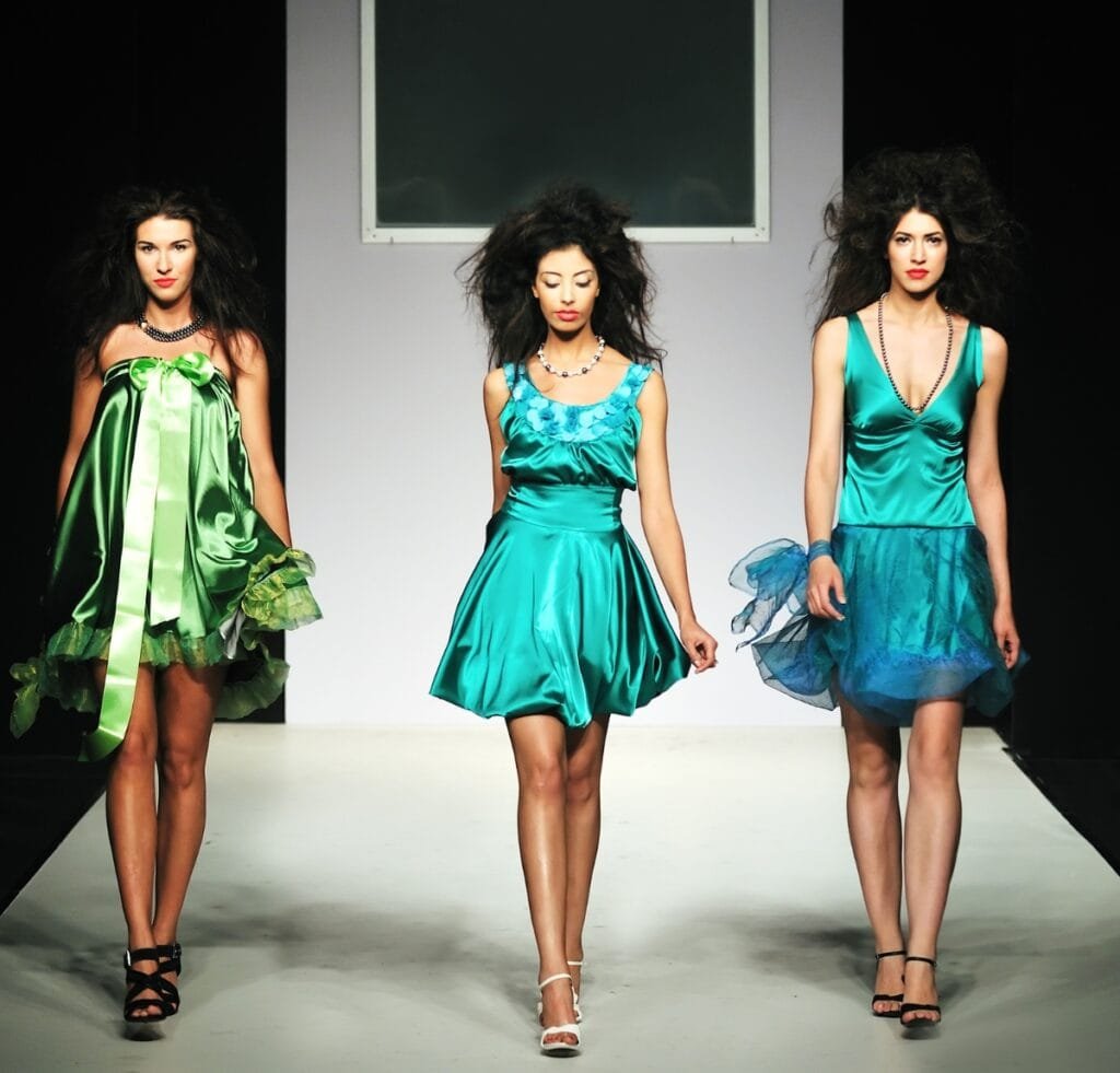 “Three female models walk a runway wearing vibrant turquoise and green satin dresses, with voluminous black hair and strappy heels against a minimalist backdrop.”