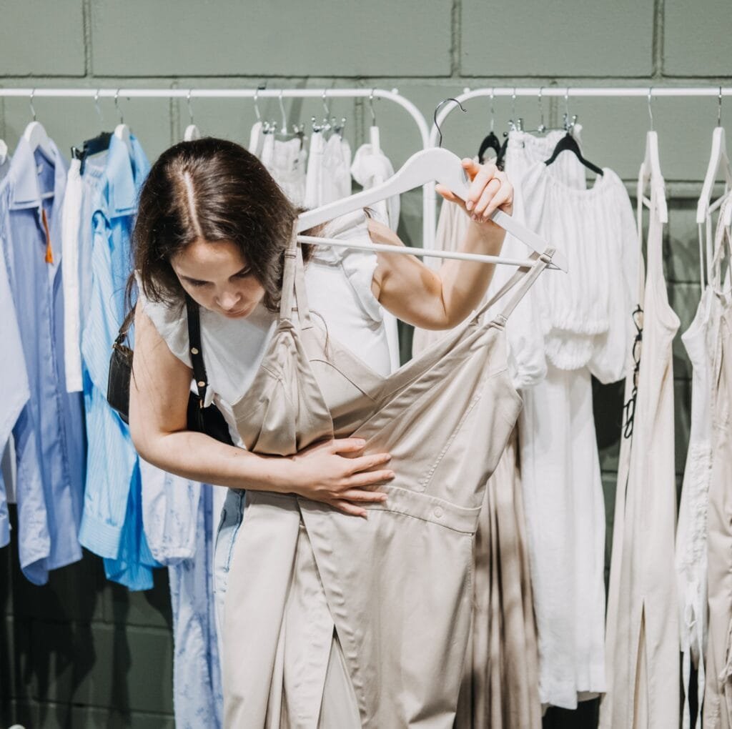 A woman in a white top browses and holds up a light beige linen dress on a rack in a boutique, surrounded by other garments hanging on white and blue hangers.