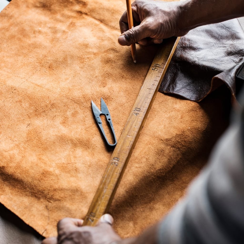 A close-up of a craftsman’s hands holding a wooden ruler and pencil over a tan buffalo leather hide, with a small cutting tool resting nearby on the leather.