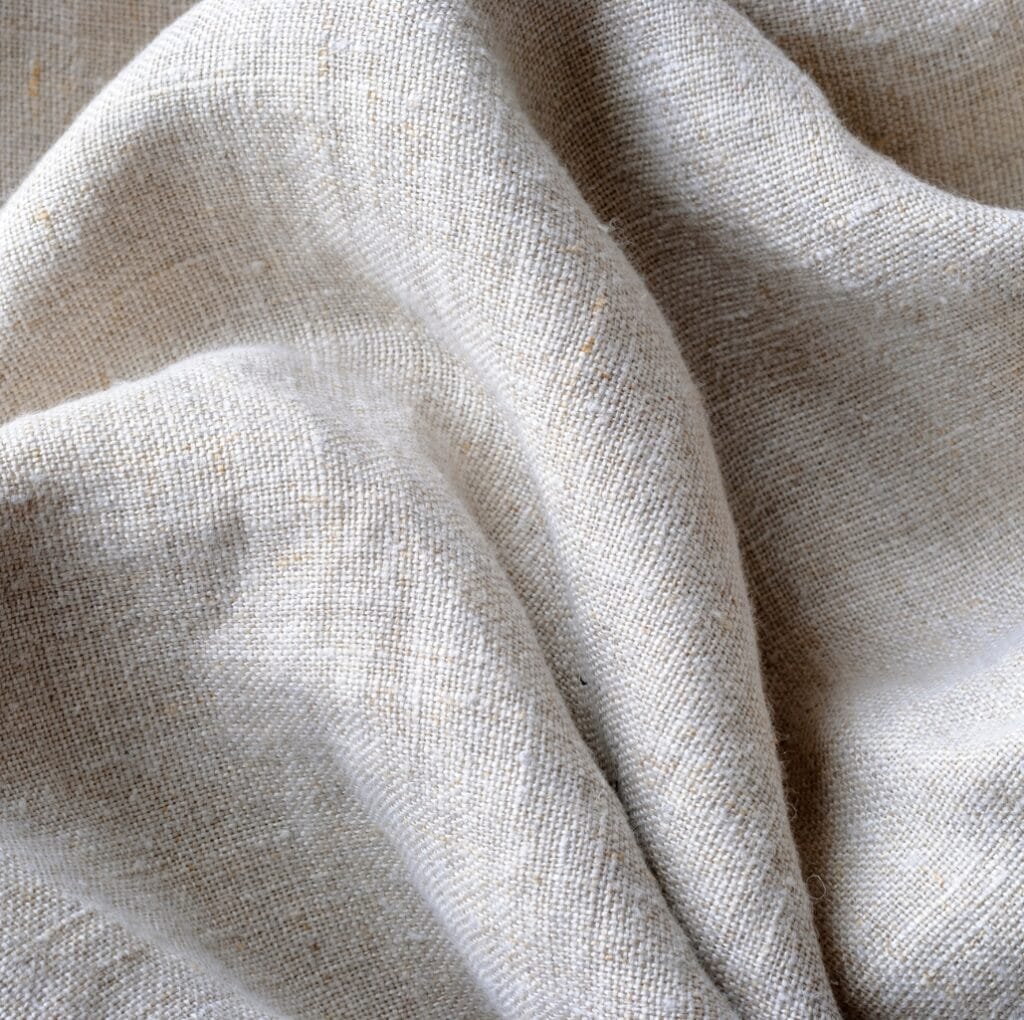 A close-up view of softly draped cambric linen fabric in a natural, light beige color, showcasing its smooth weave, delicate texture, and subtle irregularities.