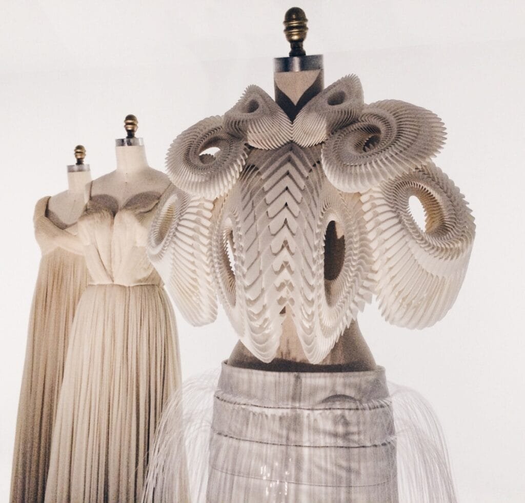 A sculptural, avant-garde dress form and two mannequins in soft beige fabrics. The central piece is an intricate, geometric couture sculpture with layered circular cutouts and pleated textures, mounted on a metallic pedestal, conveying high-fashion craftsmanship. Haute Couture Fashion