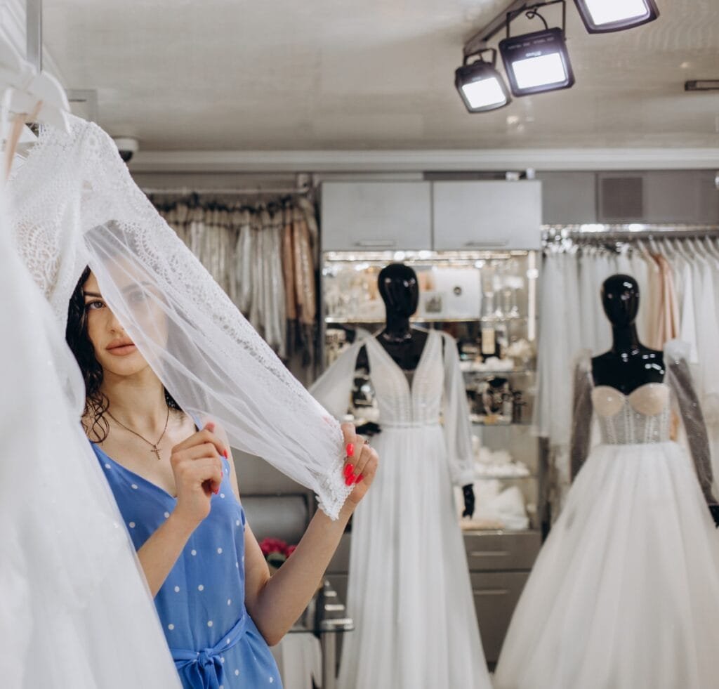 A young woman in a blue polka-dot dress examines a delicate white lace veil inside a bridal shop. She holds the veil up, partially shading her face. In the background, two mannequins wear elegant wedding gowns, and racks of dresses fill the boutique, illuminated by bright showroom lighting. Haute Couture Fashion