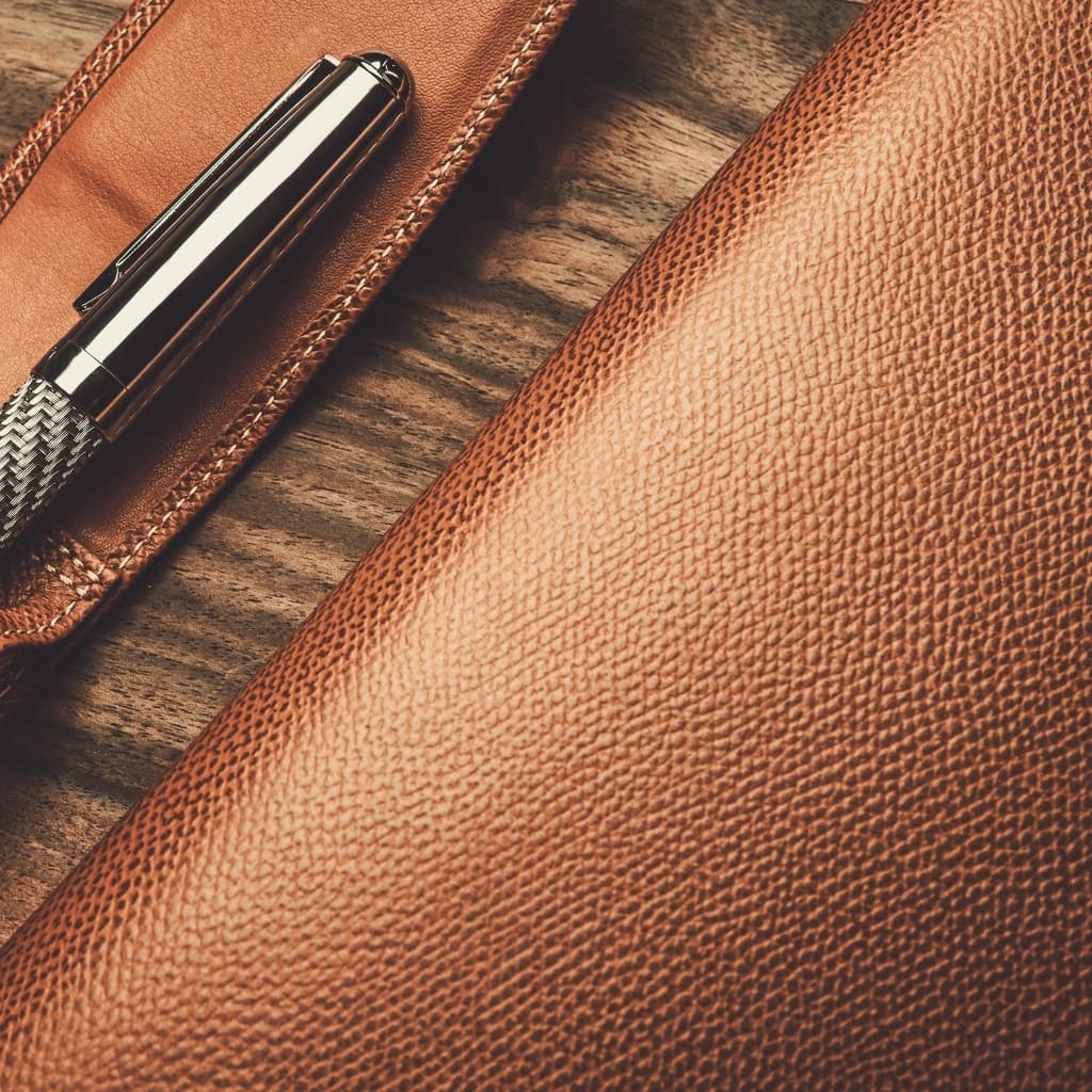 Close-up image of a textured, top grain buffalo leather surface, with a leather pen case and a shiny pen placed on a wooden surface.