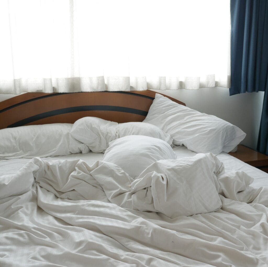 A rumpled, white bed with crumpled sheets and pillows on a wooden headboard in a sunlit room.