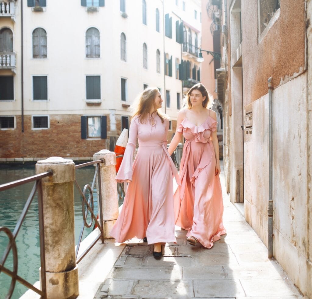 Two women walking along a sunlit canal in Venice, wearing flowing pink linen dresses. They stroll on a narrow stone walkway beside aged buildings with shuttered windows, a metal railing over the water, and gentle reflections in the canal. The scene has a timeless, romantic mood.