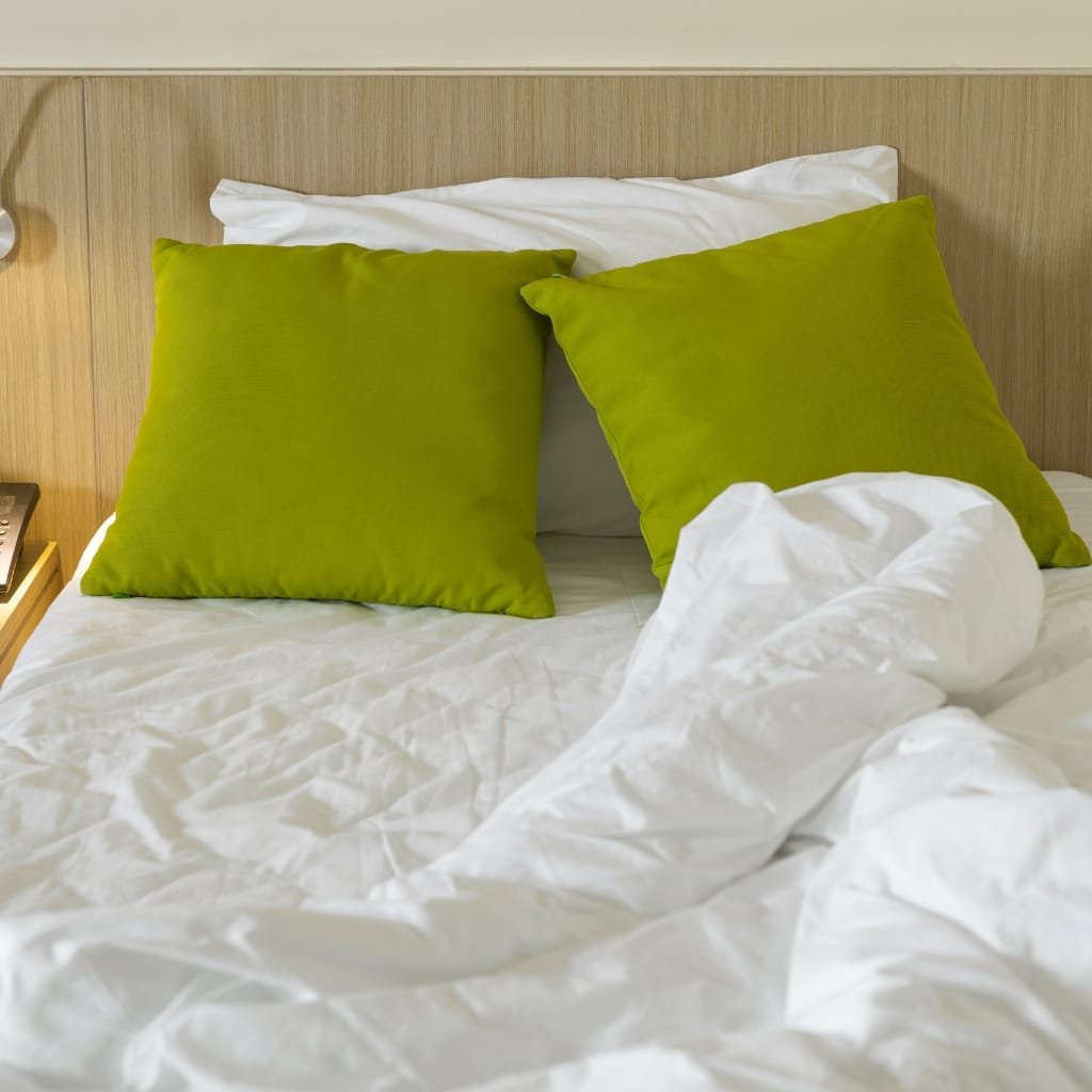 A neatly made bed with white sheets and two bright green pillows, against a light wood headboard