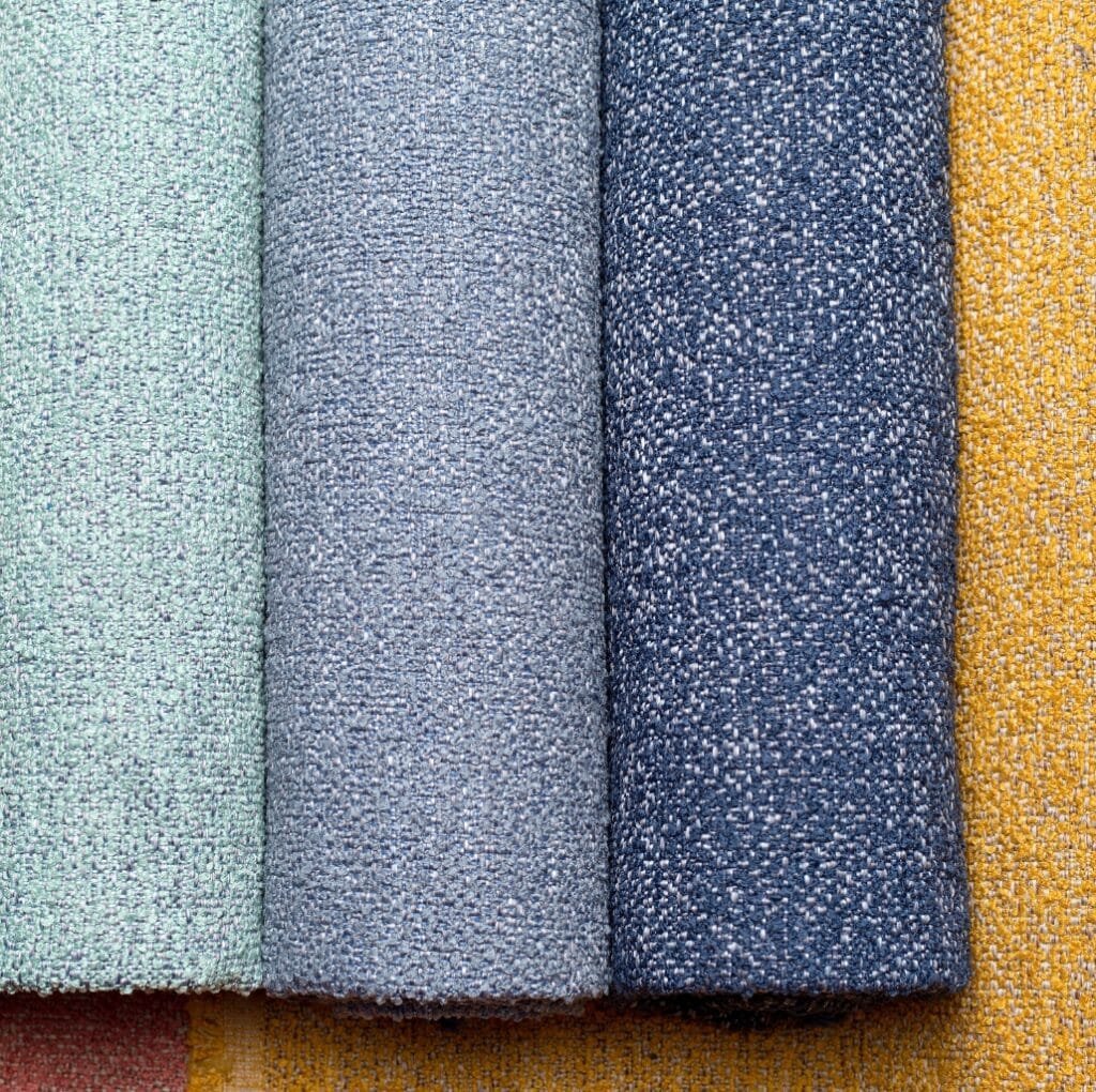 A close-up view of several rolled upholstery fabrics in soft, textured linen-like weaves. Colors from left to right include light mint green, light blue, medium denim blue, and warm mustard yellow.