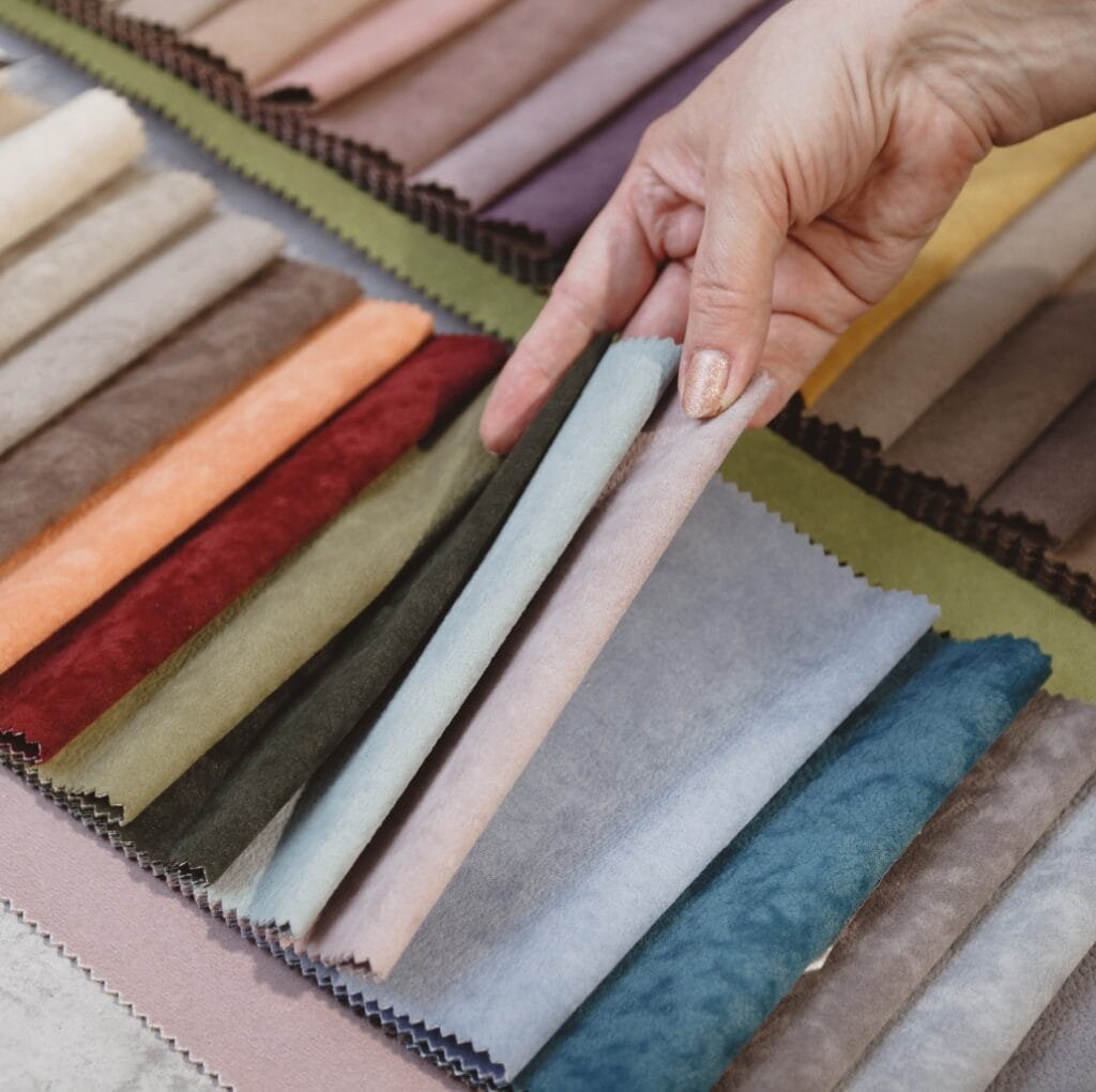 A hand holding a sample swatch from a display of upholstery fabrics in a wide range of colors, including pinks, blues, greens, and neutrals, arranged in overlapping strips with pinked edges.