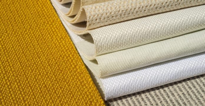 A stacked assortment of upholstery fabrics in warm neutral tones. The top left features a rich mustard-yellow textured fabric, followed by several ivory and beige woven fabrics with varying weaves, ending with a light striped beige and white material at the bottom.