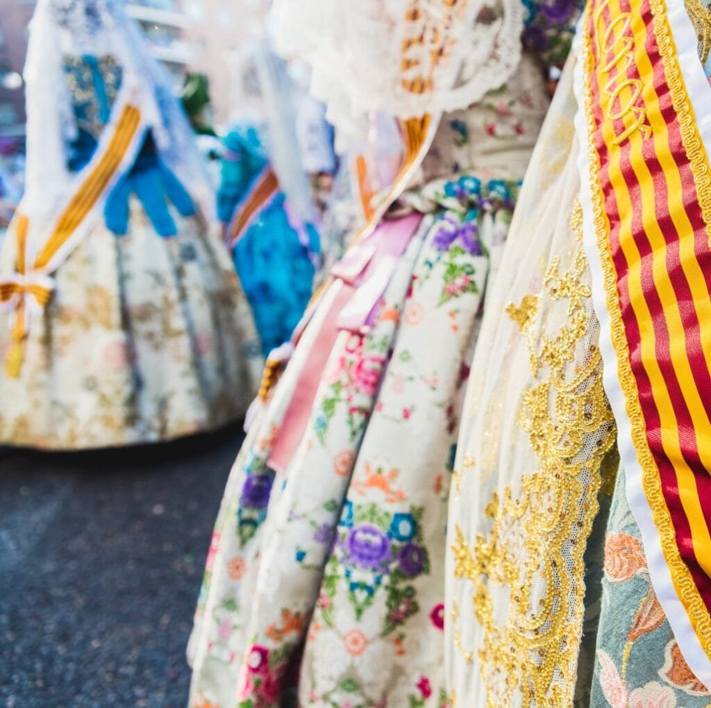 A row of beautifully patterned linen fabrics hanging outdoors. The fabrics feature floral prints in soft pastels and lace details, with bold gold embroidery and ribbon trims visible along the edges.