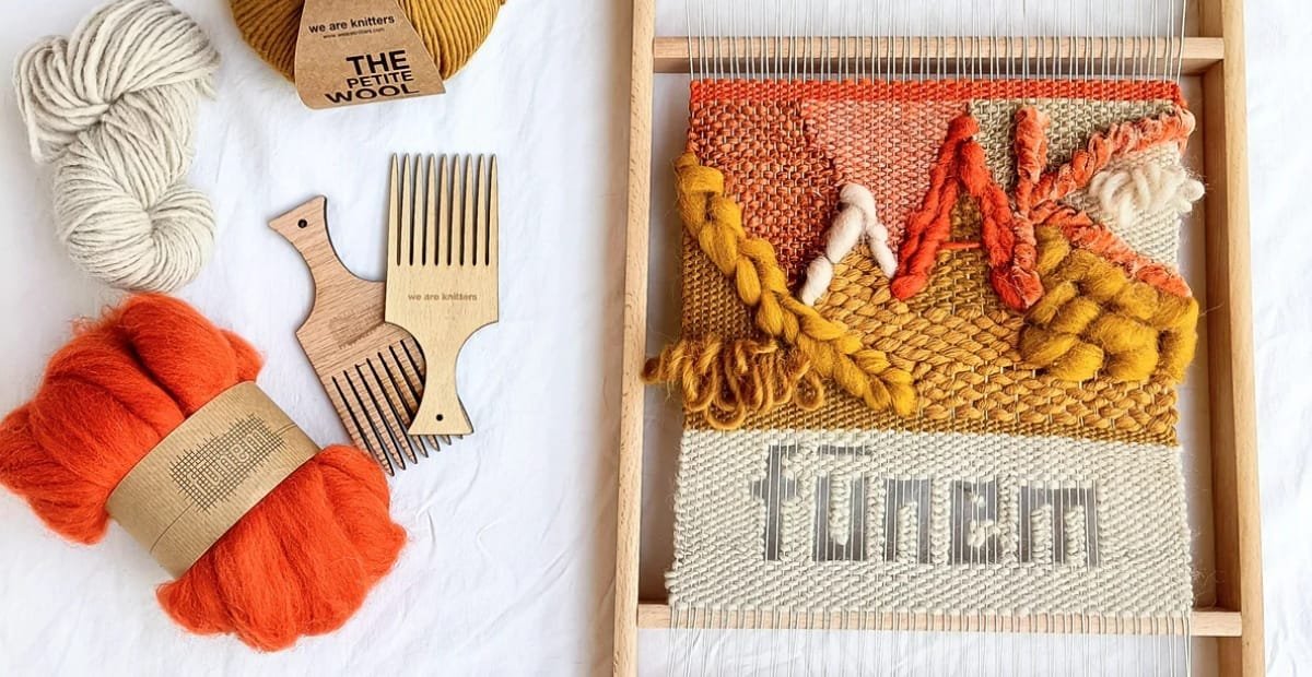 Weaving vs Knitting: A Complete Guide to Fabric-Making A framed loom weaving project with a colorful woven piece featuring warm oranges, yellows, and cream tones, plus crafting tools and yarn on the side. A skein of bright orange yarn, cream wool, and light brown yarn sit nearby, along with a wooden comb fork and a small wooden loom accessory. The woven panel in progress includes textured patterns and the word “home” or a similar word woven in gray at the bottom.