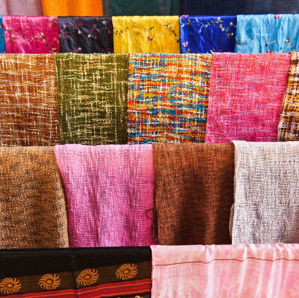 “A vibrant display of woven fabrics arranged in rows, featuring a variety of textures and colors including pinks, browns, greens, blues, and metallic threads.”
