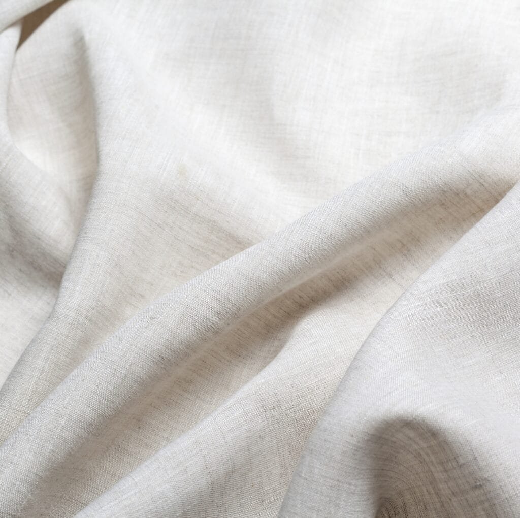 A close-up image of light beige linen fabric with soft, natural-looking texture and gentle folds.