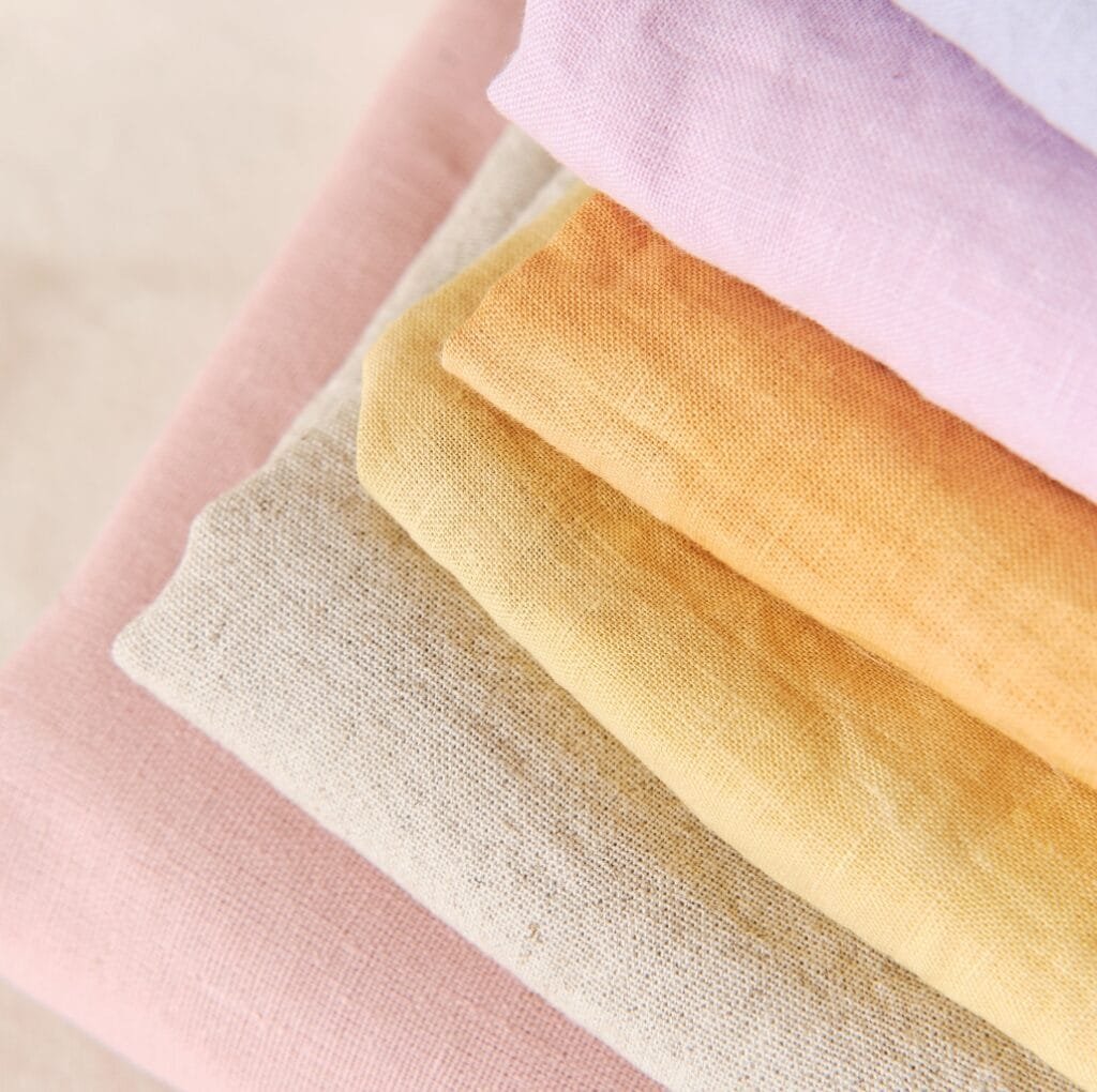 A stack of folded linen fabric in pastel colors including pink, beige, and yellow, arranged neatly on a light surface.