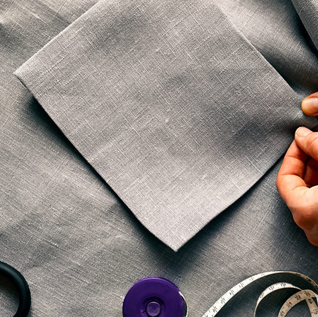 A close-up view of a light gray linen fabric being held by two hands. The textured weave is clearly visible, with a seam/corner fold and sewing details, plus a purple button and measuring tape in the periphery.