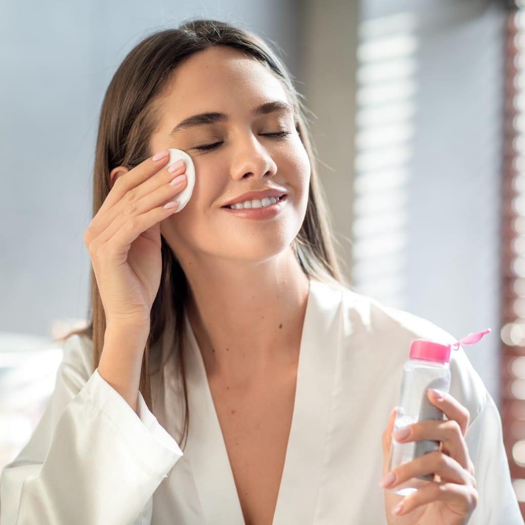 A smiling woman with long brown hair cleansing her face in a bright bathroom. She wears a white robe and gently applies a cotton pad to her cheek with her eyes closed, holding a clear bottle with a pink cap in the other hand.


