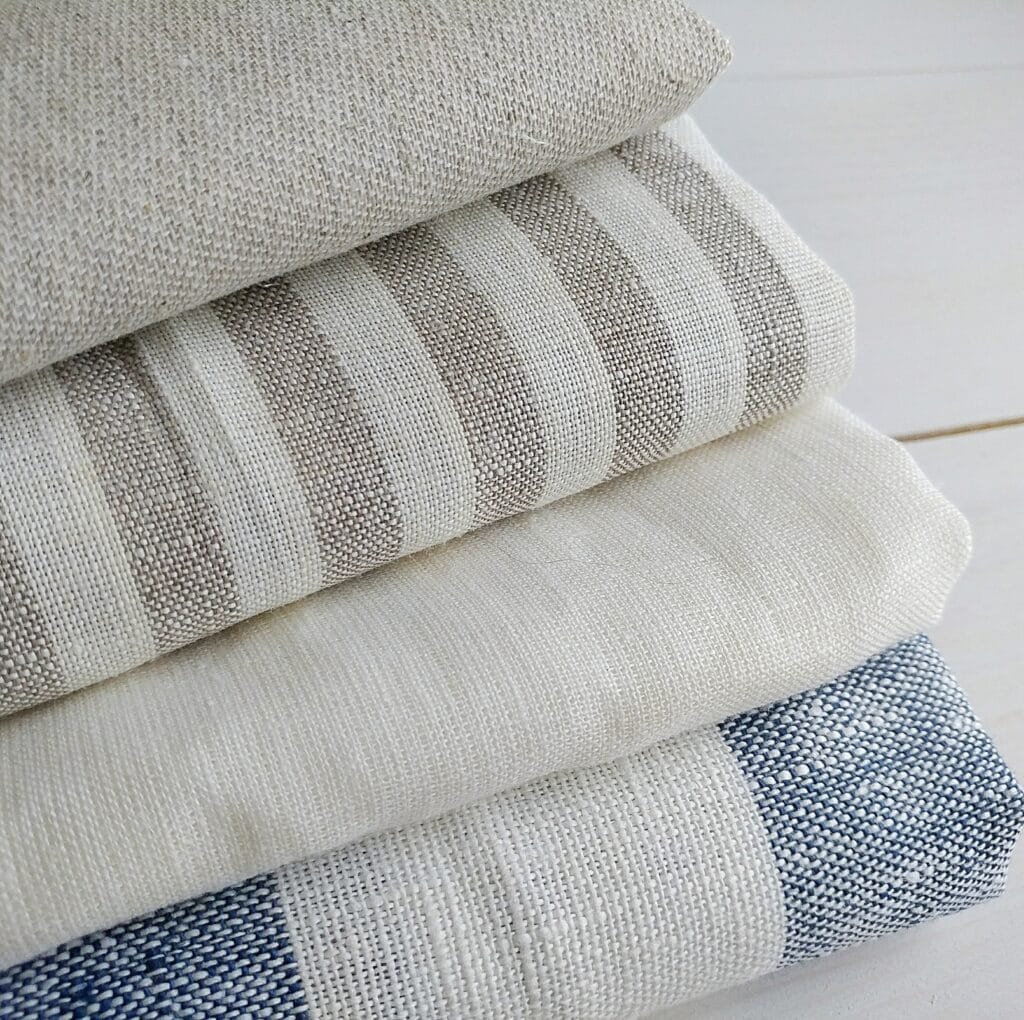 A stack of folded linen fabrics in neutral shades (beige, cream, and blue) with a visibly tight, close weave and subtle texture.