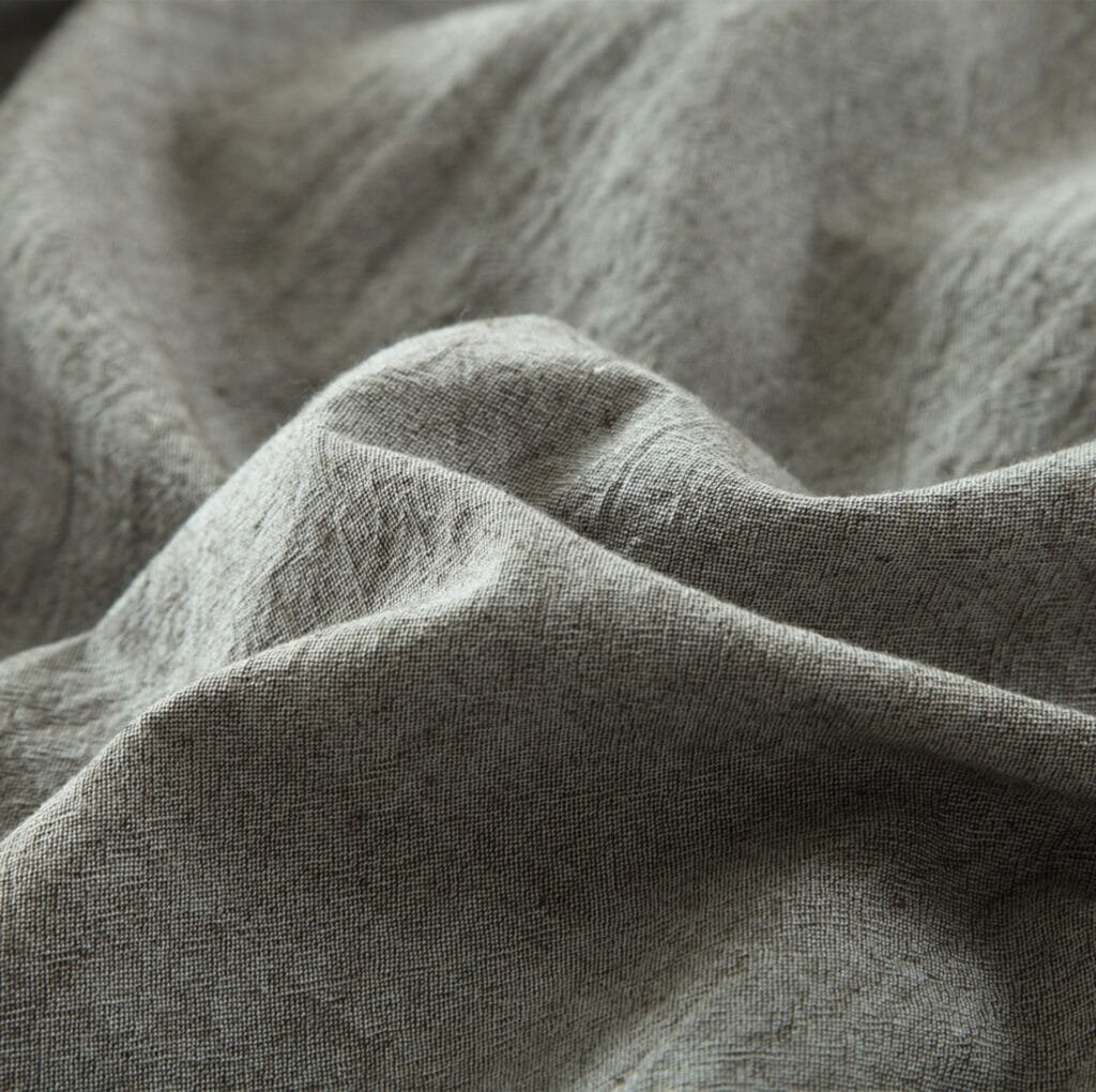 A close-up view of soft, natural linen fabric with visible weave and gentle folds, showcasing its textured, matte surface in neutral gray-beige tones.
