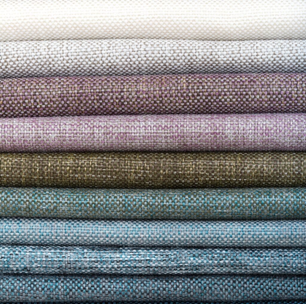 A stack of folded upholstery linen fabrics in soft, muted colors ranging from cream and beige to lavender, moss, and blue, each with a woven texture.